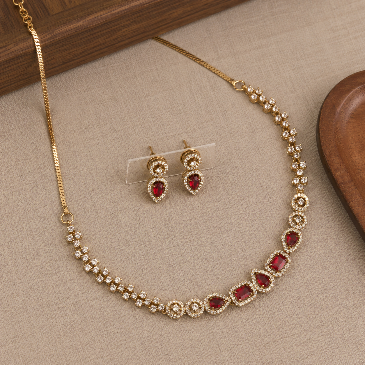 Gold-tone jewelry set with a necklace and matching earrings, featuring red gemstone accents and clear stones, displayed on a beige textured surface with wooden props.