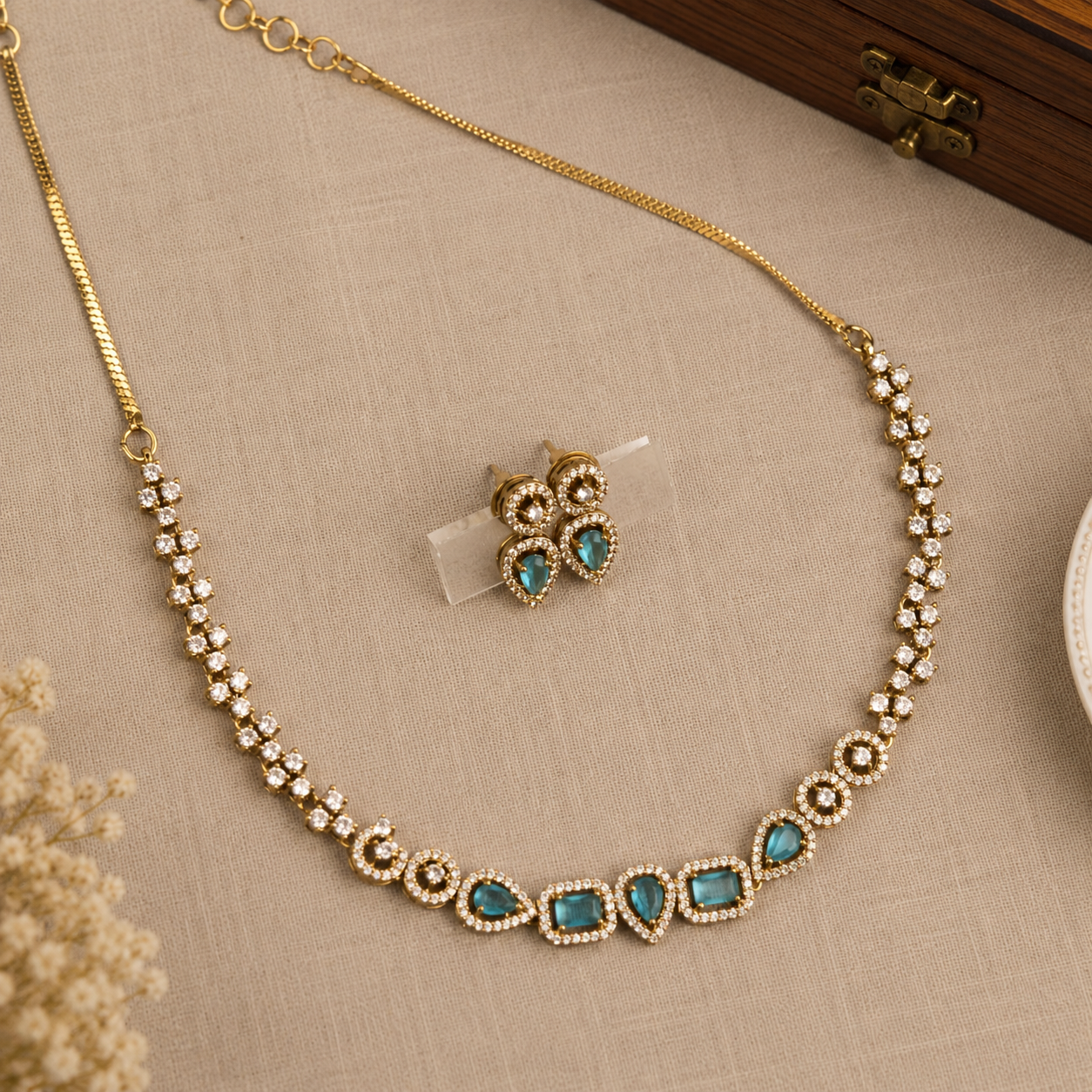 Gold-tone necklace and matching stud earrings set with turquoise blue stones and clear rhinestone accents, arranged on a beige textured fabric surface.
