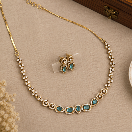 Gold-tone necklace and matching stud earrings set with turquoise blue stones and clear rhinestone accents, arranged on a beige textured fabric surface.