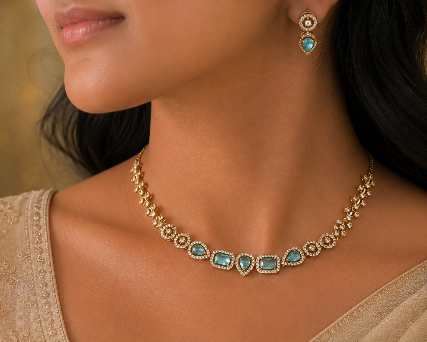 Gold necklace and matching drop earring with turquoise-blue stones, shown on a model's neck and upper chest against a softly blurred warm background.