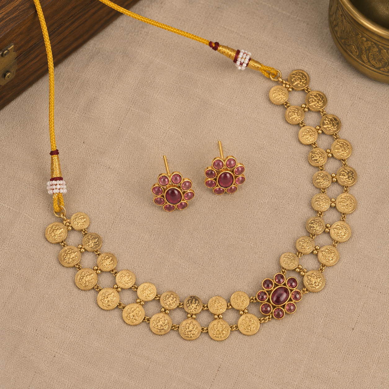 Gold coin-style necklace and matching flower-shaped earrings with red gemstone centers, displayed on a beige fabric surface.