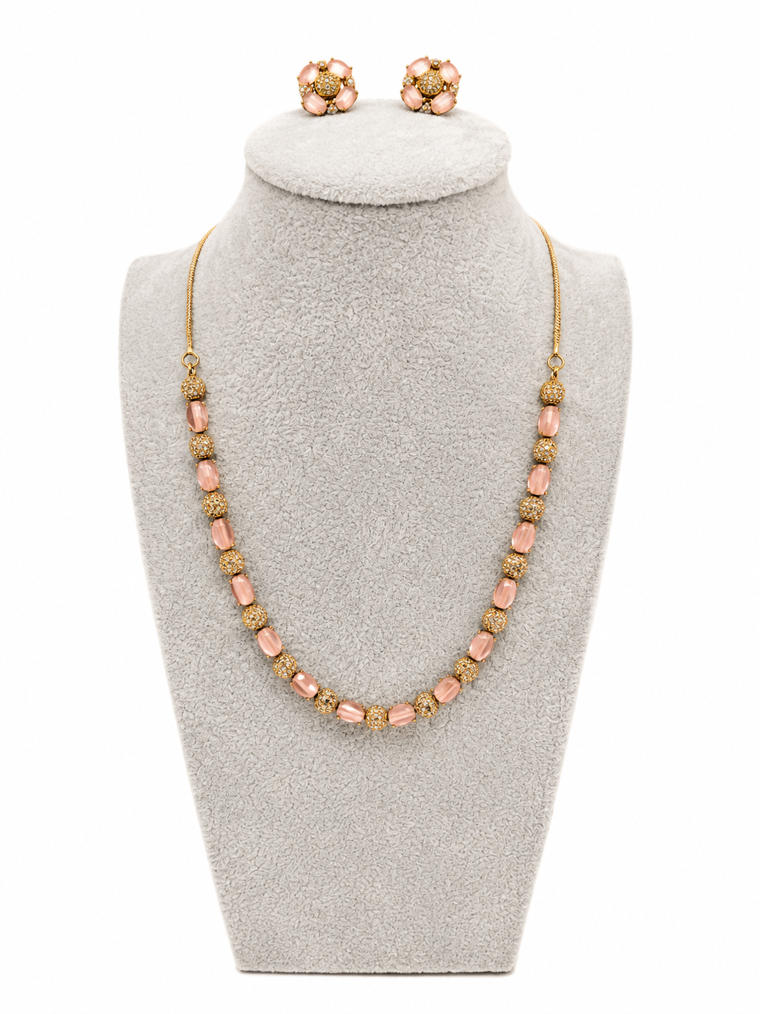 A gold-tone statement necklace and matching stud earrings displayed on a light gray jewelry bust. The necklace features alternating smooth blush pink beads and textured gold spacer beads, with matching gold-and-pink flower-shaped stud earrings positioned at the top of the bust.