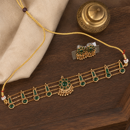 Gold-toned traditional Indian jewelry set featuring a choker necklace and matching earrings, embellished with green stones and gold filigree, displayed on a dark wooden surface with a beige cloth and brass decorative object.