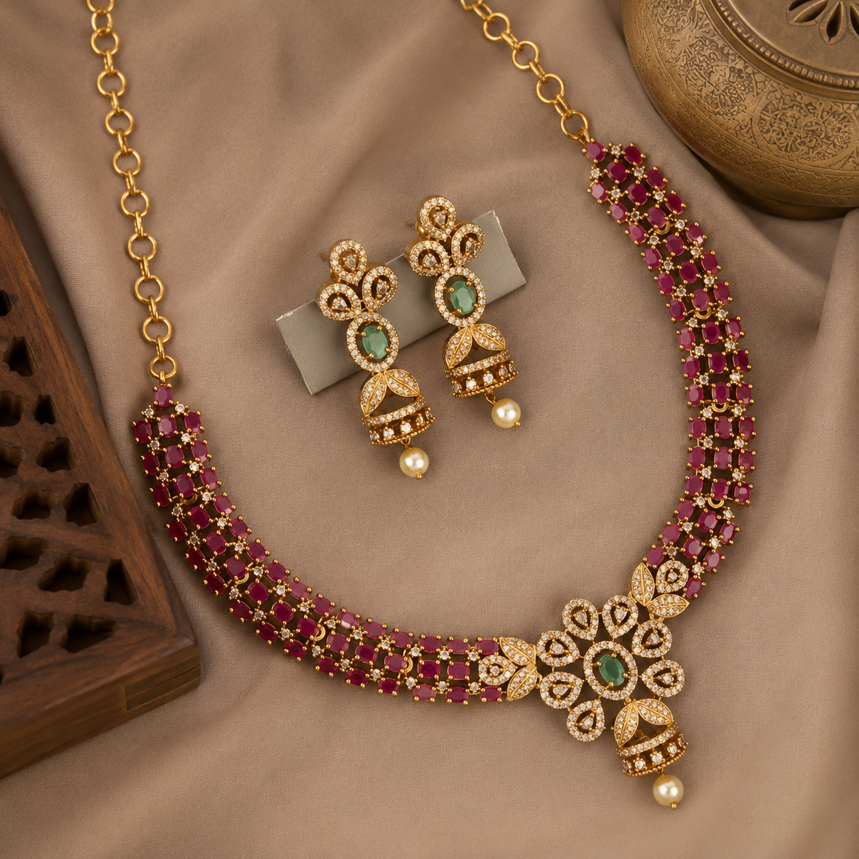 Gold-plated traditional Indian jewelry set featuring a necklace and matching earrings, embellished with deep red stones, small clear stones, and green gemstones, displayed on a beige fabric background.
