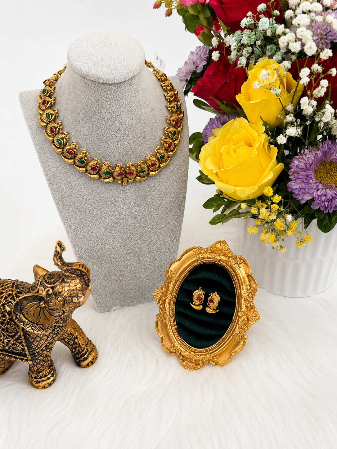 A gold-toned statement necklace with red and green stone accents displayed on a gray velvet mannequin bust, alongside a small ornate gold mirror, a decorative elephant figurine, and a colorful flower arrangement.