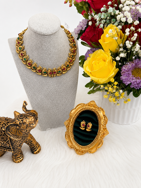 A gold-toned statement necklace with red and green stone accents displayed on a gray velvet mannequin bust, alongside a small ornate gold mirror, a decorative elephant figurine, and a colorful flower arrangement.