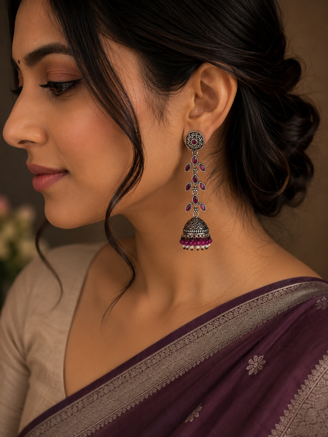 A woman in profile wearing a dangling silver-toned jhumka earring with purple and white stone accents, paired with a deep purple saree.