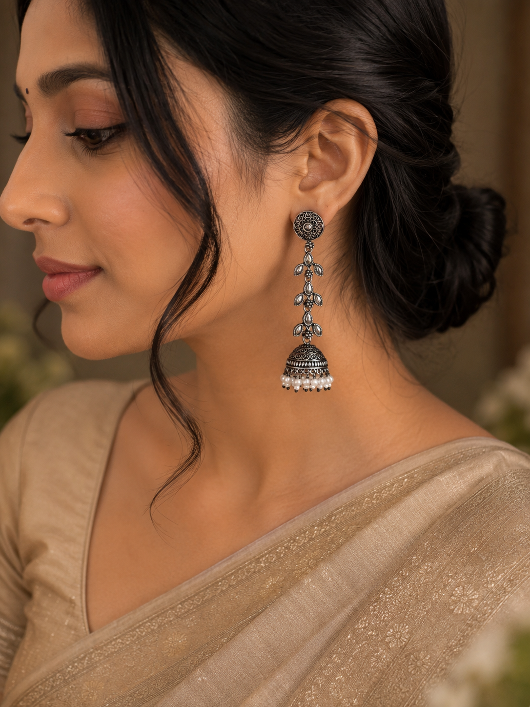 A close-up side profile of a woman wearing a traditional Indian jhumka earring. The earring is made of silver-toned metal with intricate filigree work and a dangling bell-shaped charm finished with small white stones.