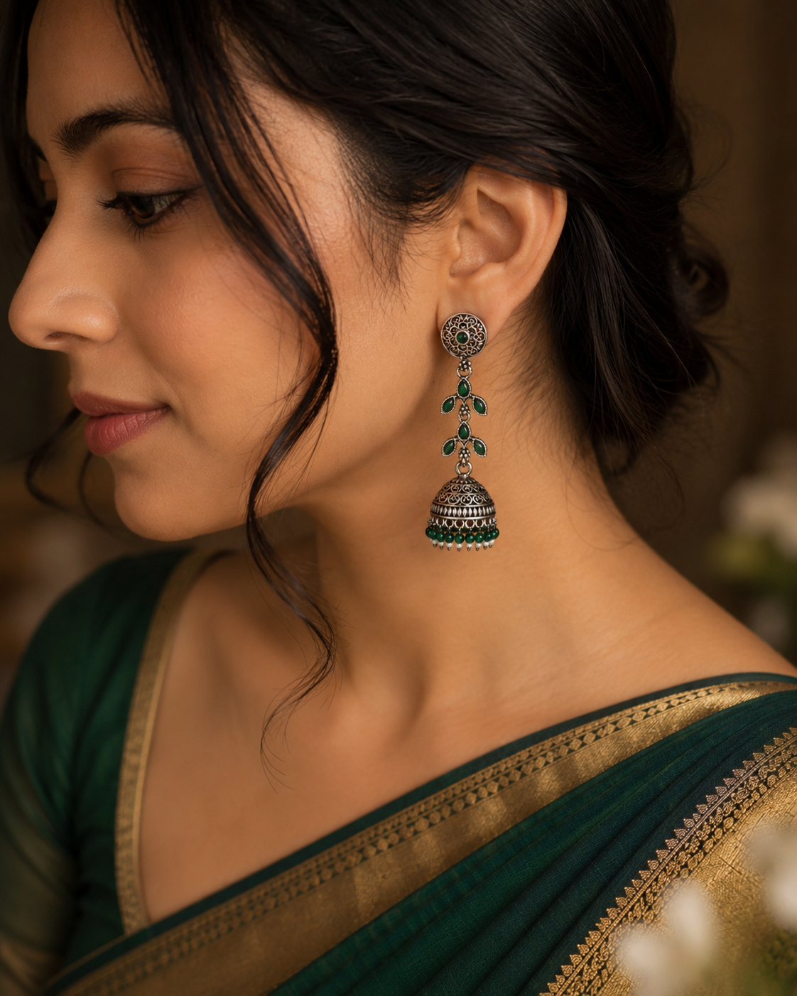 A close-up side profile of a woman wearing a traditional Indian jhumka earring. The earring features a silver-toned filigree bell-shaped dangle with green stone accents.