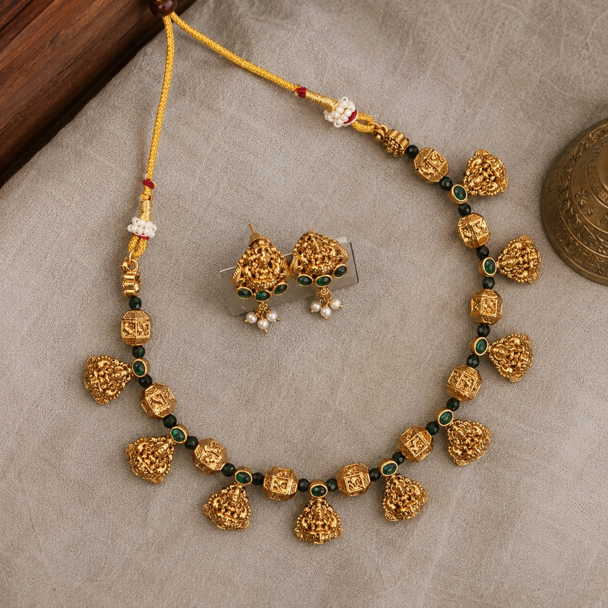 Gold-toned traditional Indian jewelry set featuring a necklace and matching earrings, decorated with textured gold beads, small green stone accents, and white pearl-like drops, displayed on a neutral fabric background.