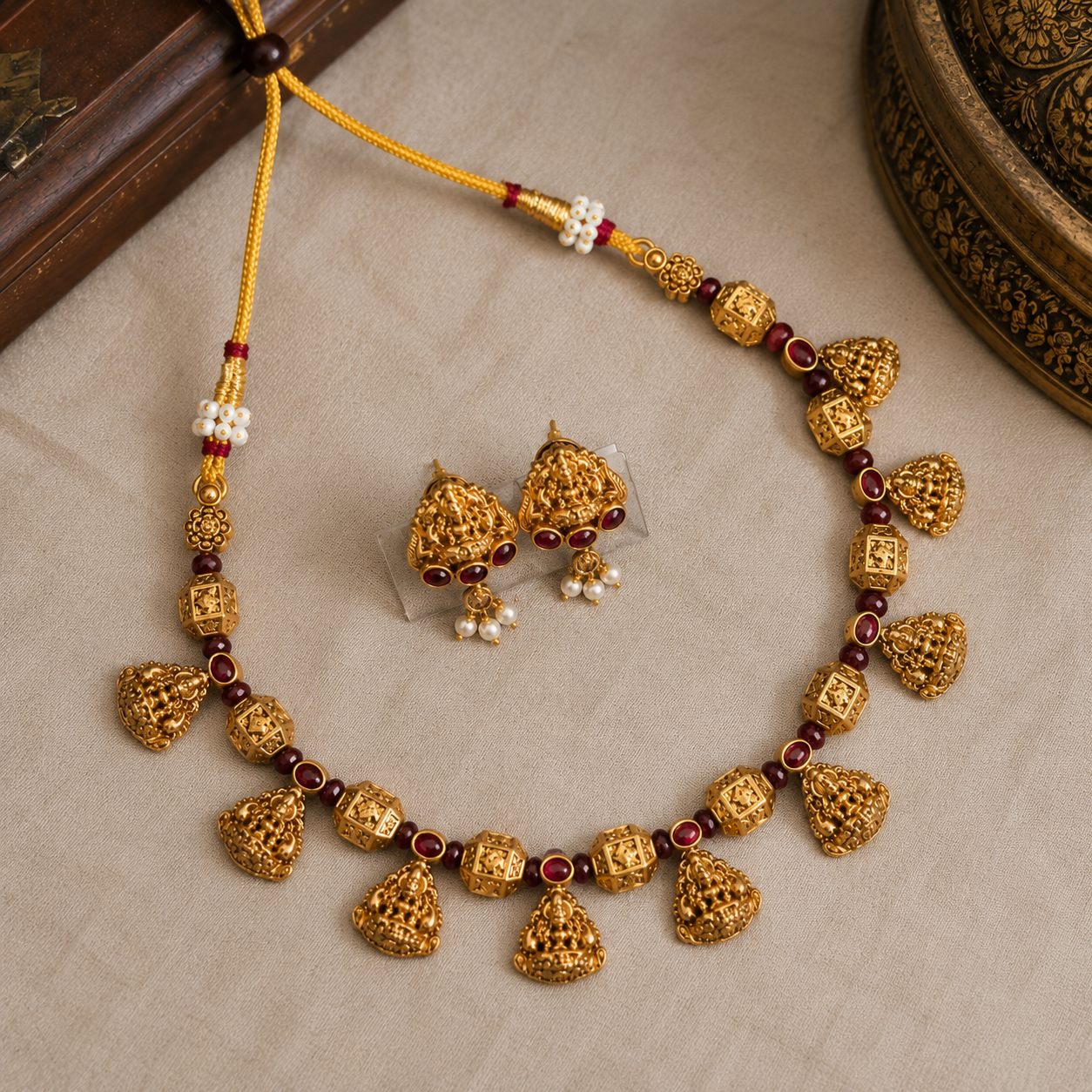 Gold-toned traditional jewelry set featuring a necklace and matching earrings with ornate filigree bell-shaped charms, red gemstone accents, and small pearl details, arranged on a light beige fabric surface.