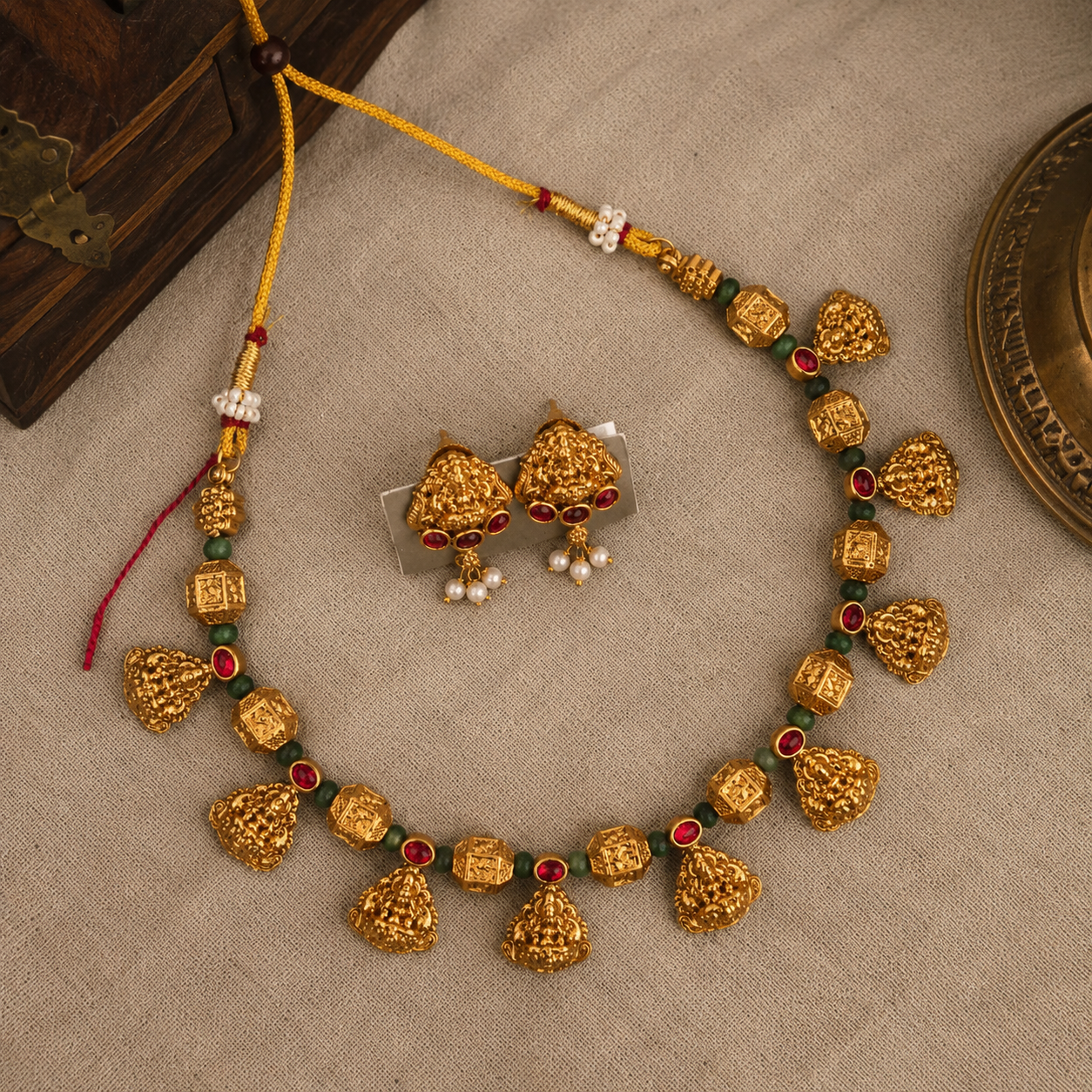 Gold-toned traditional necklace and matching earrings with red and green stone accents and white pearl-like beads, arranged on a beige fabric surface.