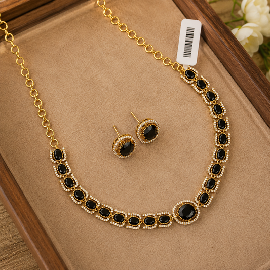 Gold-tone necklace and matching stud earrings set with oval black stones and small clear crystal-like accents, displayed on a tan jewelry tray inside a wooden box.