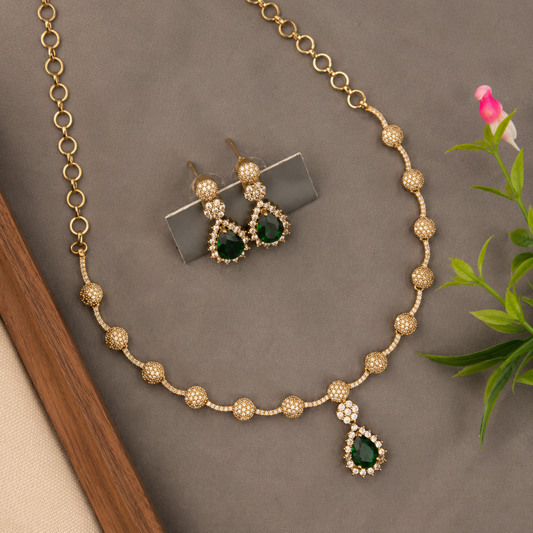 Gold-tone necklace and matching earrings set with deep green teardrop-shaped stones and small clear rhinestone accents, arranged on a gray surface with decorative leaves and a wooden edge.