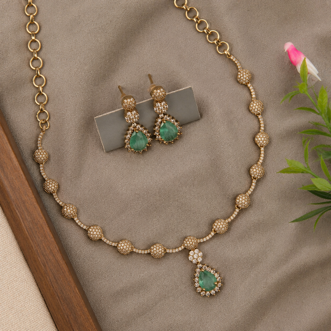 Gold-toned traditional necklace and matching drop earrings with green teardrop-shaped stones and small clear stones, arranged on a beige fabric surface with decorative leaves and a wooden edge.