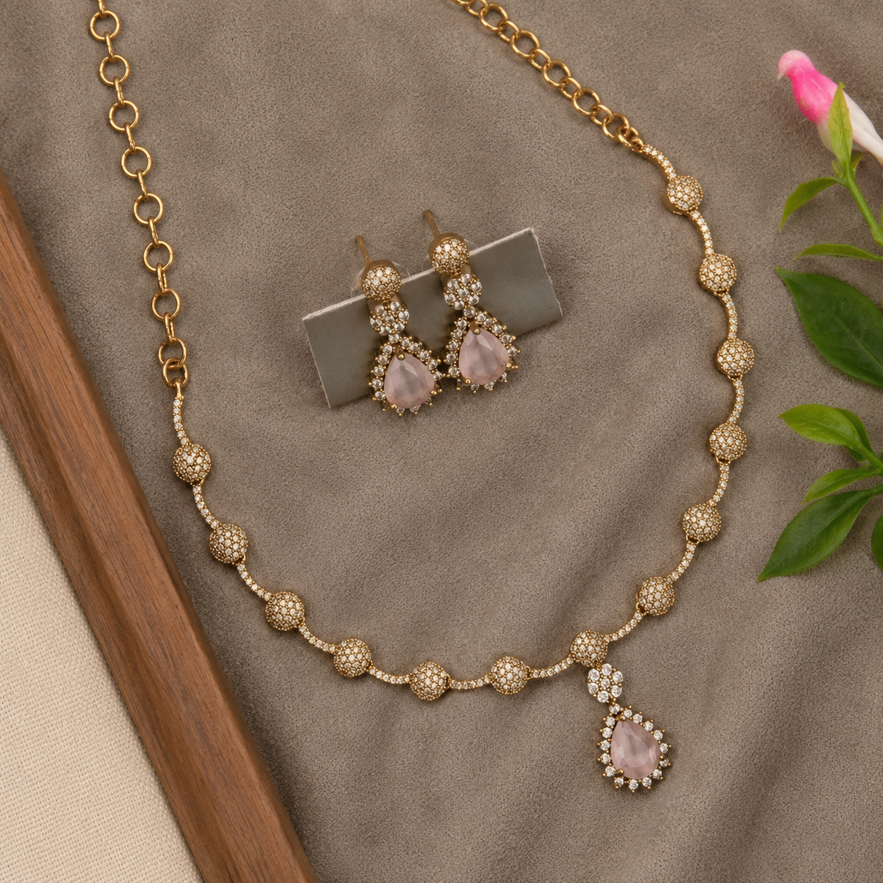 Gold-toned jewelry set featuring a beaded necklace and matching drop earrings with pale pink teardrop-shaped stones, displayed on a textured taupe fabric surface with decorative leaves and a flower.