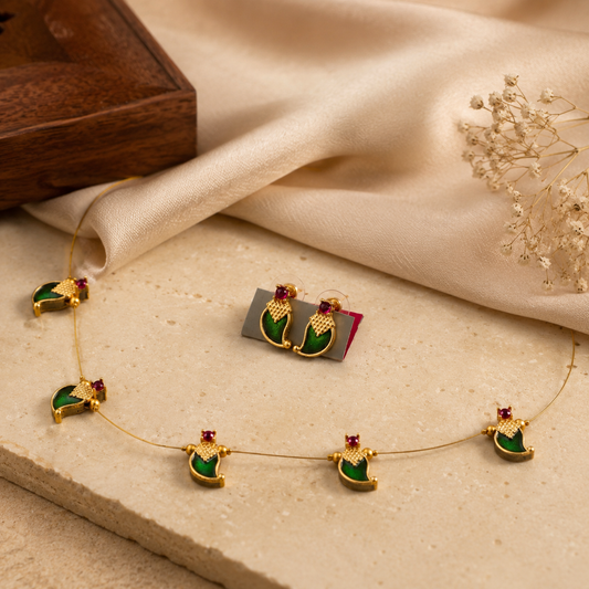 Gold-toned jewelry set featuring crescent-shaped earrings and a matching necklace with green enamel details and small red gemstone accents, arranged on a beige stone surface with cream satin fabric and dried floral decor.