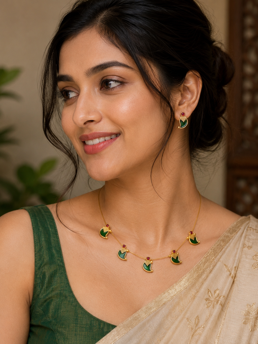 A woman wearing a gold crescent moon necklace and matching drop earrings with green enamel details, styled with a dark green blouse and a beige saree.