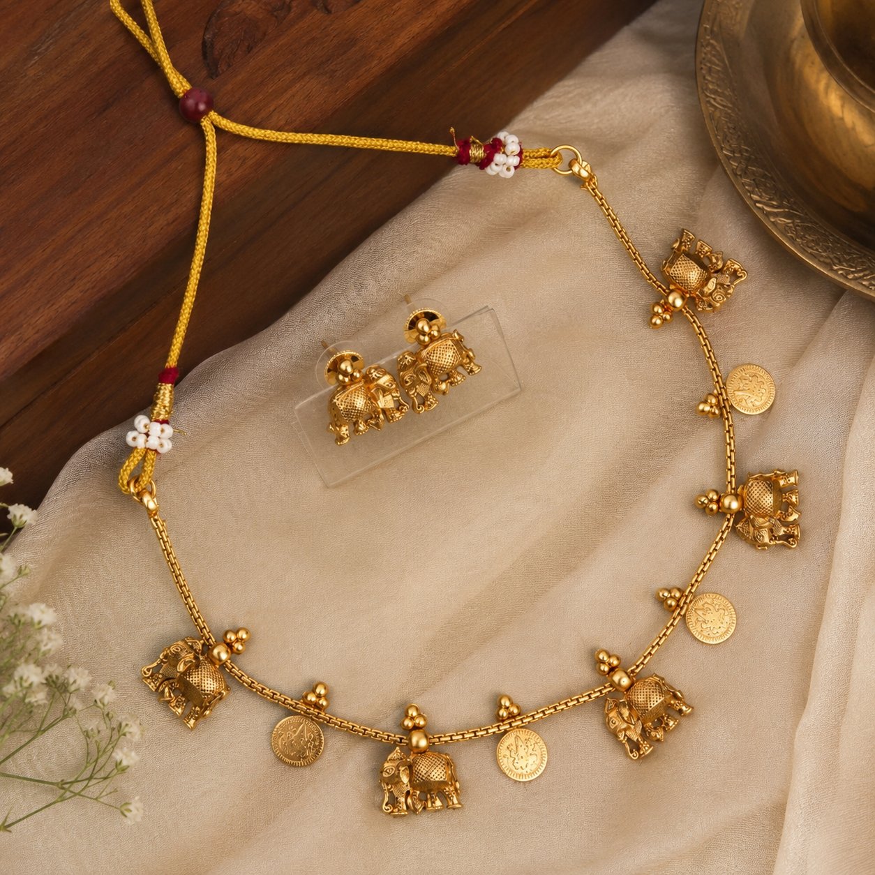 Gold-toned necklace and matching earrings featuring elephant motifs and coin details, displayed on a beige fabric surface with a wooden tabletop and decorative props.