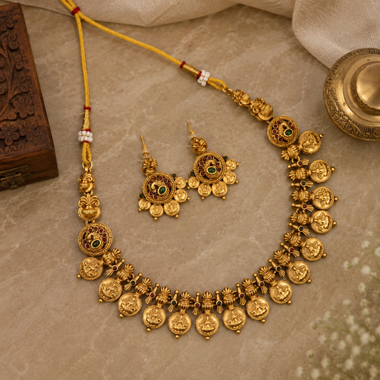 An ornate gold-toned jewelry set laid out on a beige stone surface, including a necklace with dangling round motifs, matching drop earrings, and a yellow cord. The set is styled with decorative props such as a carved wooden box, a brass vessel, and draped fabric.
