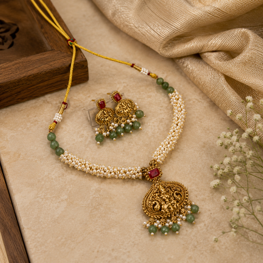 An ornate traditional Indian jewelry set featuring a necklace and matching earrings. The necklace has a double-strand pearl-beaded chain with green bead accents, a gold jhumka pendant with a red stone, and a large detailed gold pendant.