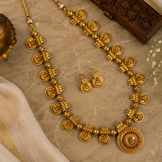 A traditional Indian gold necklace with matching earrings, featuring detailed temple-style motifs and a round pendant with red and green stone accents, displayed on a beige stone surface with decorative props.