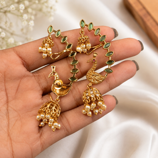 Gold-toned traditional Indian earrings displayed on a person's hand, featuring peacock and jhumka motifs with green stone accents and pearl-like bead clusters.