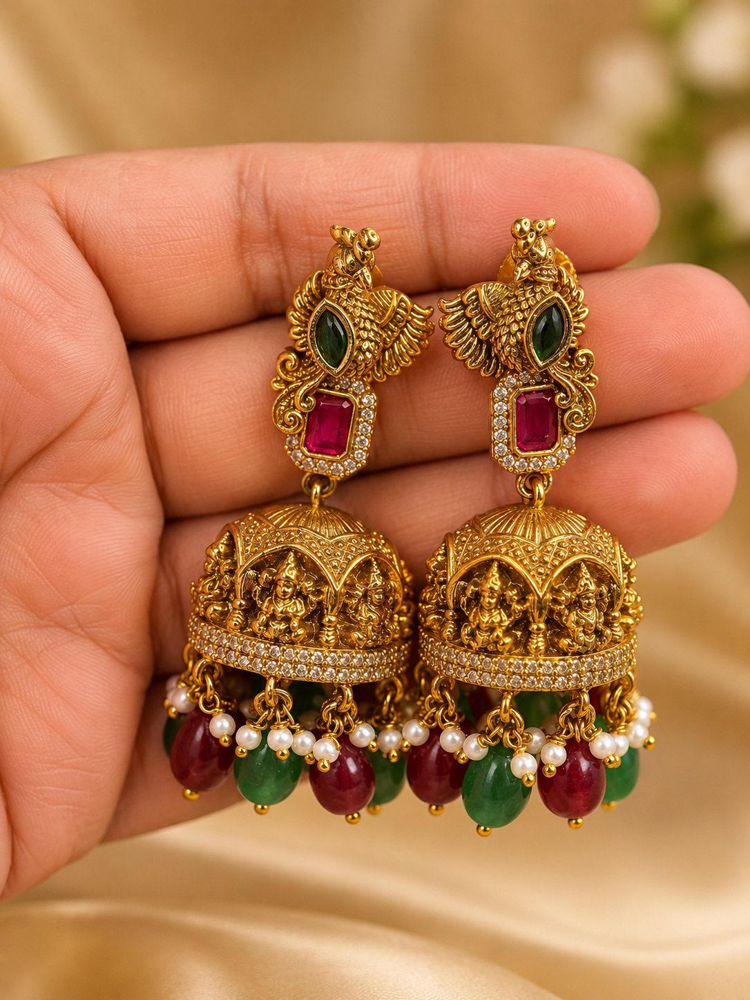 A pair of ornate gold-toned Indian jhumka earrings with green and red gemstone accents, held in a hand against a soft beige satin background.