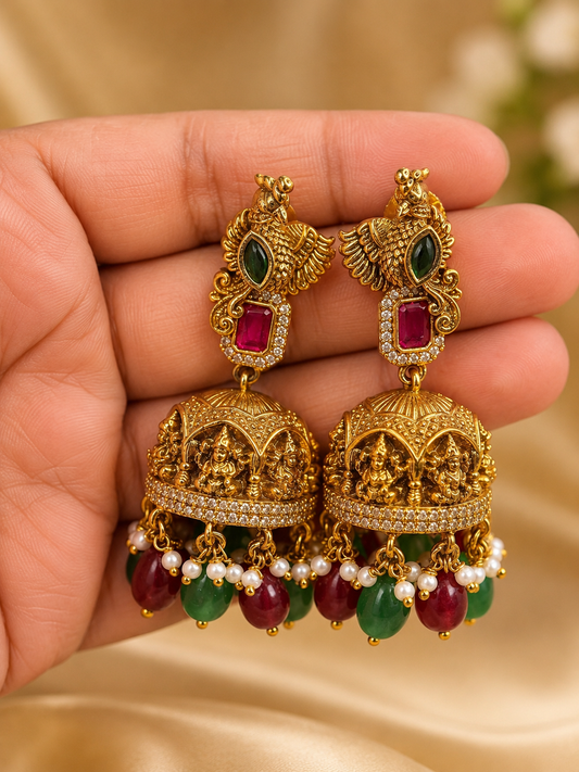 A pair of ornate gold-toned Indian jhumka earrings with green and red gemstone accents, held in a hand against a soft beige satin background.