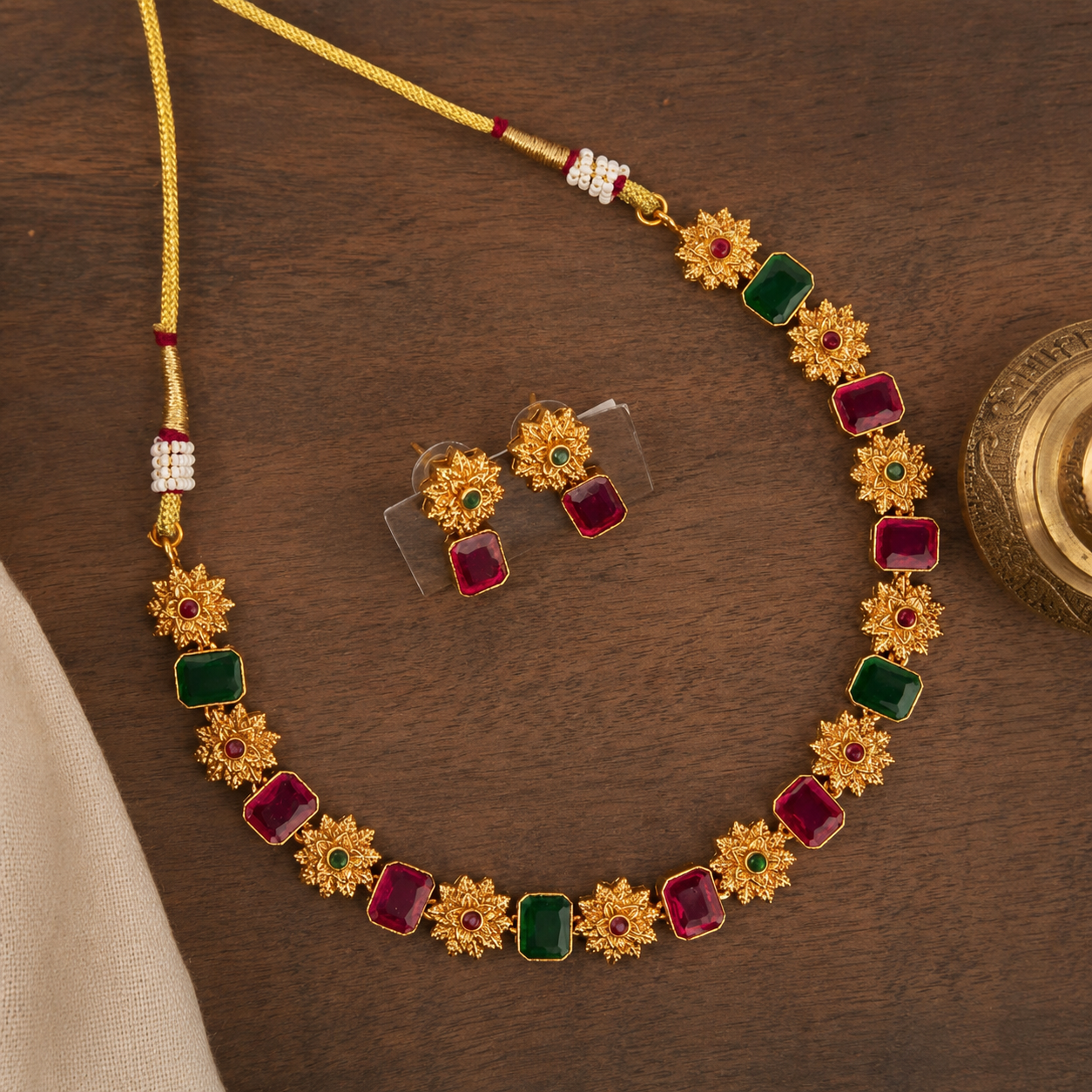Gold-toned necklace and matching earrings with alternating red and green rectangular stones and floral starburst motifs, displayed on a dark wooden surface.