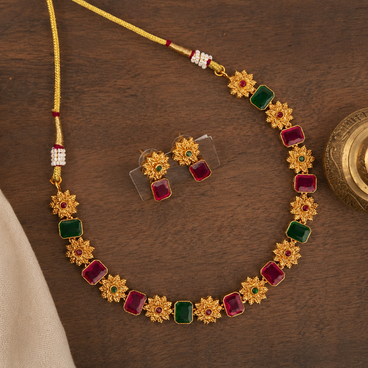Gold-toned necklace and matching earrings with alternating red and green rectangular stones and floral starburst motifs, displayed on a dark wooden surface.